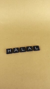 Black letter tiles spelling out the word halal on a light brown paper background, representing Islamic dietary law, permissible food and drinks, and religious certification for muslims