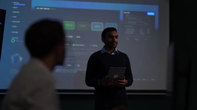A listener in the foreground of a dark room comes into focus as the speaker at the screen behind him falls away into shallow depth at an AI company in Bangkok, Thailand.