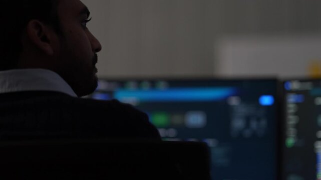A bearded engineer in half profile studies his monitors in near darkness, the interface reflected across the side of his face inside a dimly lit machine learning startup office.