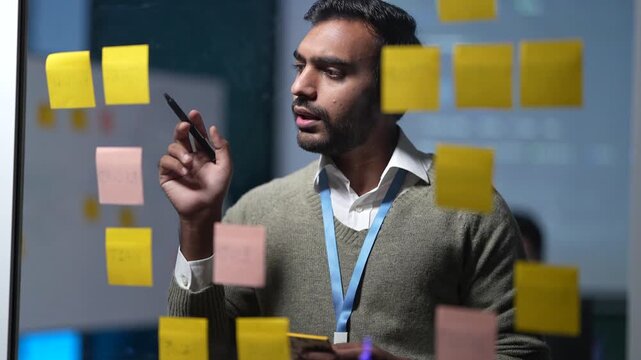 An engineer behind a note covered glass wall holds a hand to his chin, then lifts a pen to mark the step that has been bothering him inside a dark AI startup office.