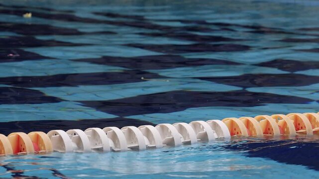 Swimming pool lane divider floats on water surface. Orange and white plastic floats form a line. Blue water ripples gently around the divider. Ideal for fitness, sports, or aquatic activity scenes