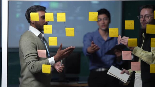 A team member pointing across the board directs his colleagues to the card that has to move before anything downstream can run inside a dimly lit machine learning startup office.