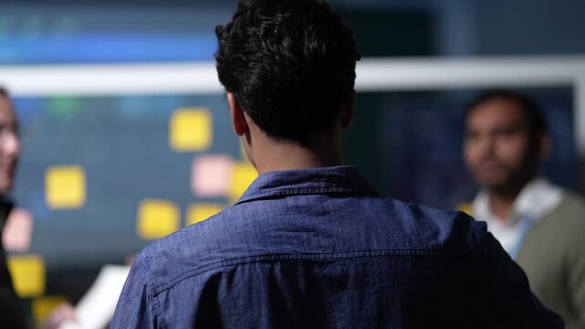 An over the shoulder view of a planning session holds a developer's back in frame while his teammates work the notes on the far side inside a dark AI startup office.