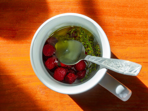 White mug of herbal tea with red berries and wild thyme on sunlit wooden table