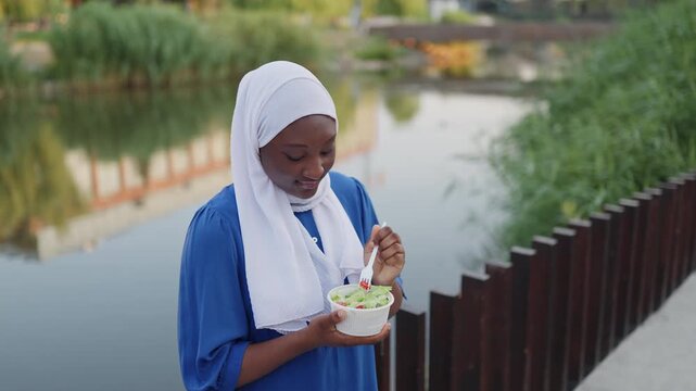 A young African American Muslim woman wearing a white hijab enjoys a healthy salad by a tranquil lake