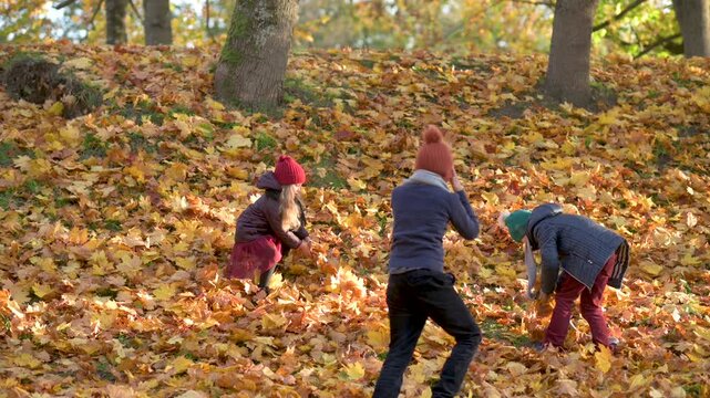 hree happy kids, two boys and a girl, playing together in autumn park, picking up and throwing colorful fallen leaves.