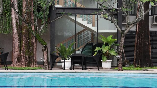 Modern house with poolside seating and lush green trees. Deep blue water reflects the sky above. Black furniture invites relaxation by the pool. Large windows frame the open, airy interior
