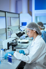 A focused female scientist in protective gear conducts medical research using a microscope and test...