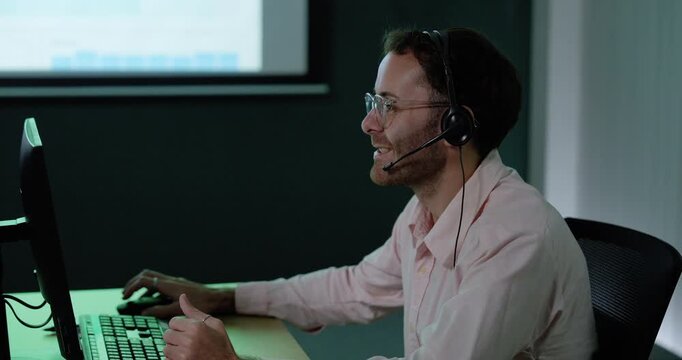 Adult male customer support professional wears headset, types at computer and gestures while speaking, smiling and handling digital communication during call center service work in modern office.
