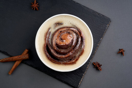 Cinnamon roll bun with sweet glaze in a paper takeout bowl on a black slate board with cinnamon sticks.