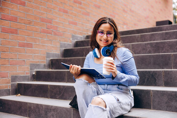 Happy student learning with coffee and headphones on campus stairs