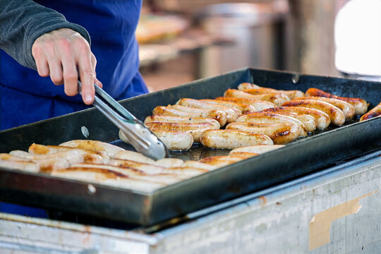 Turning Sizzling Bratwurst Sausages with Tongs on an Outdoor Flat Top Grill