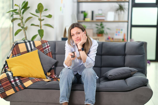 Bored businesswoman watches television while sitting on office sofa. Professional rests face on hand pointing remote control to screen during break