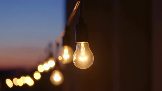 Warm hanging light bulb glows in sharp focus beside a dark wall as a diagonal string of softly blurred lights recedes toward a blue and pink dusk sky in the background