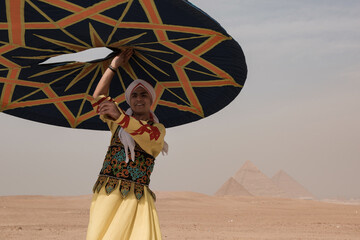 Smiling Egyptian tanoura folk dance performer raises geometric decorated disk overhead before hazy Giza Pyramids in vibrant traditional costume ideal © .shock