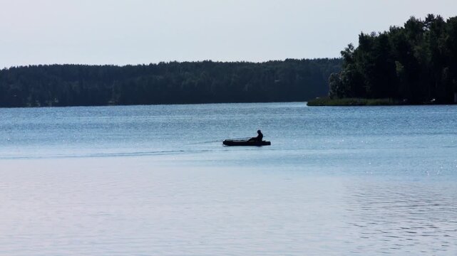 Silhouette of person fishing from inflatable boat on calm, sparkling lake water with distant lush green forest shoreline under bright, clear sky, emphasizing tranquility.