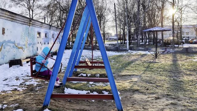 Young child swings on playground swing set during winter day with snow on ground and bare trees, sunlight illuminates scene with playful atmosphere
