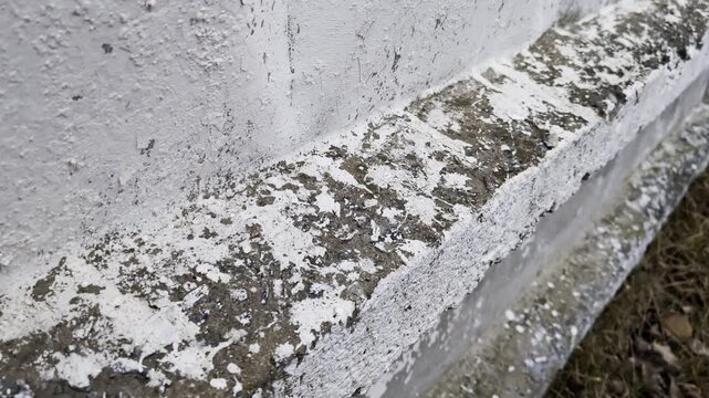 Weathered concrete surface with peeling white paint shows texture and material decay. Close-up detail of rough, old, crumbling facade, exterior building structure.