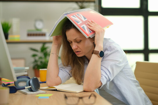 Stressed woman holds English workbook over head while sitting at office desk. Learner reacts with frustration pausing study during difficult task