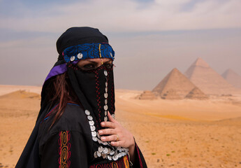 Veiled Egyptian woman wearing ornate traditional niqab with silver coin ornaments and embroidered black robe stands at Giza plateau with iconic © .shock