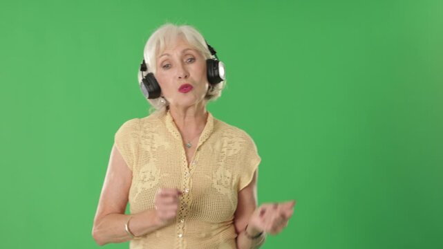 Elderly woman stands against green screen, wearing headphones, moving rhythmically and dancing while enjoying music 