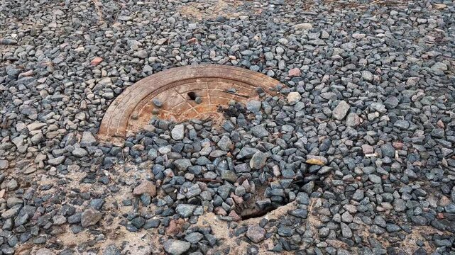 Rusted circular metal drainage cover partially submerged in grey gravel and sandy soil, showing texture of coarse stones and a small opening in earth below