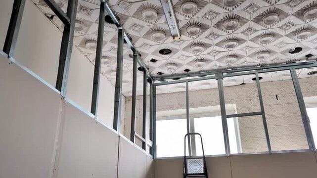 Metal stud framework construction with gypsum board panel installation in commercial building interior. Unfinished room renovation showing decorative ceiling tiles and fluorescent light fixture.