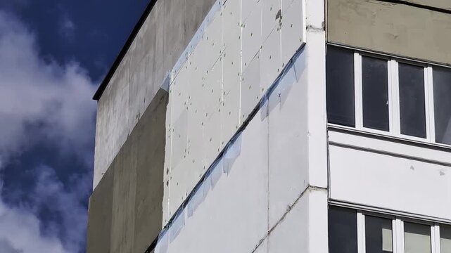 Building facade renovation process with insulation panels installation on residential apartment block exterior, showing modern energy efficiency upgrade work on multi-story structure under blue sky.