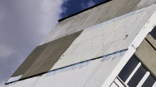 Building facade renovation process, partial installation of exterior insulation and cladding panels. Multiple construction stages visible, concrete wall section, white insulation, existing windows