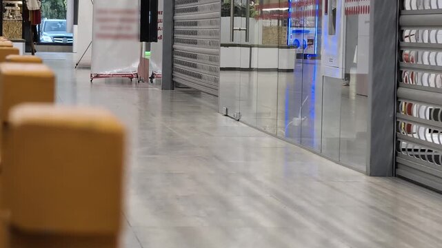 Empty commercial hallway with closed roller shutters of stores and reflections on glass facades, blurry yellow bollards in foreground, quiet atmosphere during evening or closure