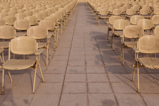 Symmetrical rows of weathered cream metal chairs receding to a vanishing point along a central stone aisle at a sunlit open air outdoor auditorium