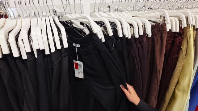 Woman browses selection of pants on hangers, choosing black trousers from clothing display in retail store, showing range of apparel colors