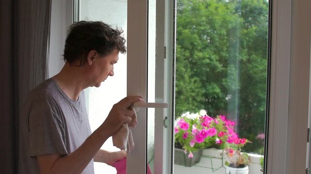 A man is washing windows in his apartment. He uses a spray bottle and a cloth to make the glass clean