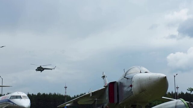 Military jet aircraft on static display at aviation museum, powerful warplane on ground, helicopter flying in distance above exhibits, cloudy sky background, other aircraft parts visible outdoors.