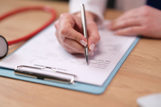 Medical professional fills printed form near stethoscope on desk. Doctor hand writes patient information completing clinical medical history with pen