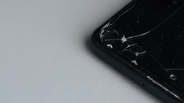 Close-up cracked screen protector on black smartphone corner background. Damaged mobile phone protector glass, severe damage, technology accident, repair concept