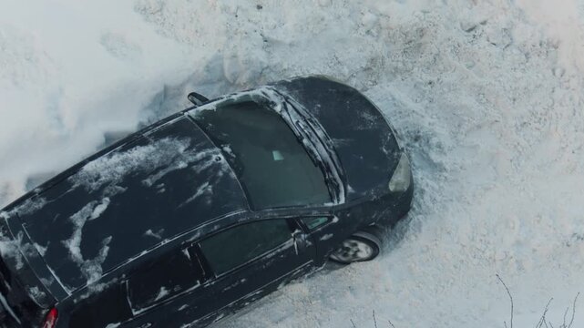 Two men pushing black car stuck in deep snow drift on cold winter day, vehicle wheels spinning, struggling to move forward on snowy road, slow motion