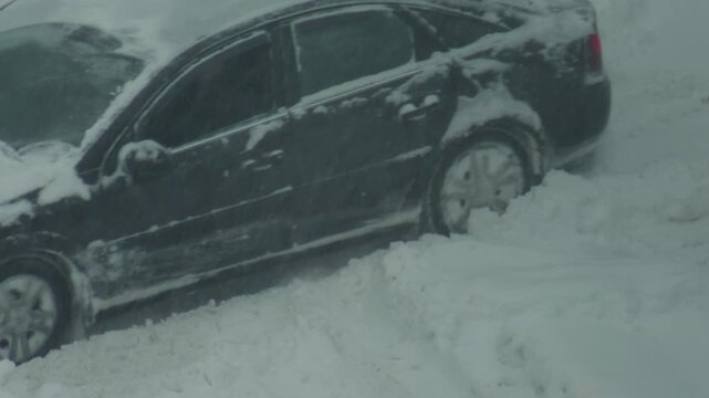 Automobile stuck in deep snowdrift during winter blizzard condition, front wheel spinning unable to gain traction, challenging road situation, slow motion