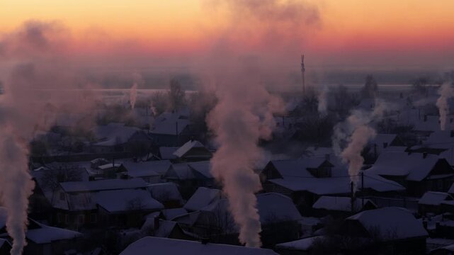 Winter frosty morning cityscape aerial view with smoke plumes rising from chimneys of snow-covered village houses against vivid sunrise sky.