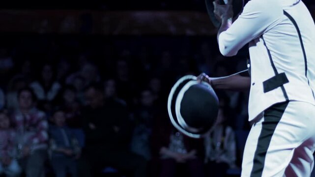 Professional male performer juggling bowler hats with expertise during circus show. Audience watches in dark venue. Entertainer displays skill and showmanship with props slow motion