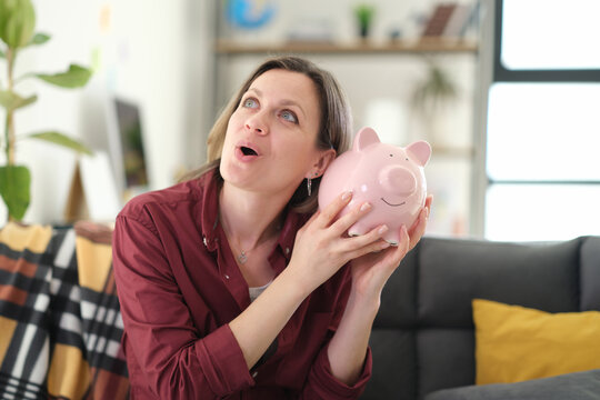Pleasantly surprised woman shakes charming pig-shaped piggy bank. Sparkling eyes and radiant expression of blonde female reveal genuine happiness