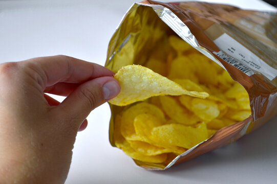 Person reaching into an open metallic bag and pulling out a single golden crispy potato chip as a savory snack on a light background