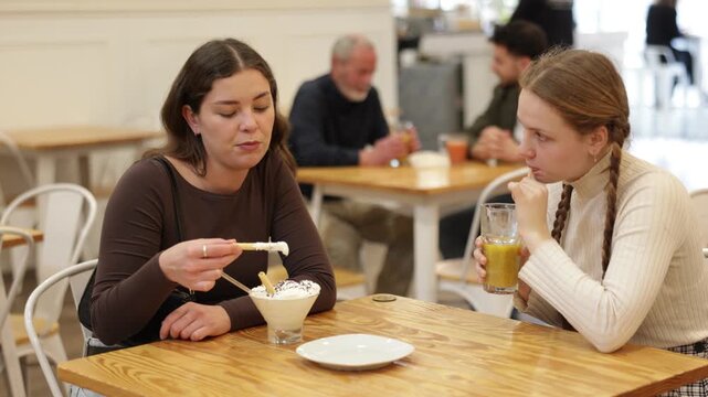 Two positive girls chatting over juice and eat ice cream while sitting at small table in cafeteria . High quality 4k footage