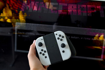 Fototapeta premium Hand holding a white Nintendo Switch OLED controller in front of a vibrant Minecraft title screen glowing on a dark television