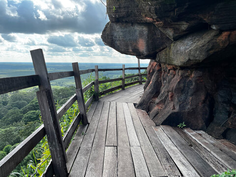 Wooden walkway around the steep sandstone cliff at Wat Phu Tok temple. Scenic mountain path trail with a wooden fence overlooking the green tropical forest and cloudy sky in Thailand