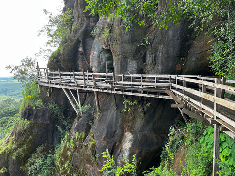 Wooden walkway around the steep sandstone cliff at Wat Phu Tok temple. Scenic mountain path trail with a wooden fence overlooking the green tropical forest and cloudy sky in Thailand