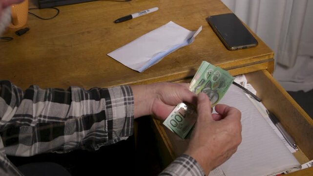 Elderly man pulls green banknotes from a cluttered desk drawer and slips the cash into a plain white envelope beside his phone.