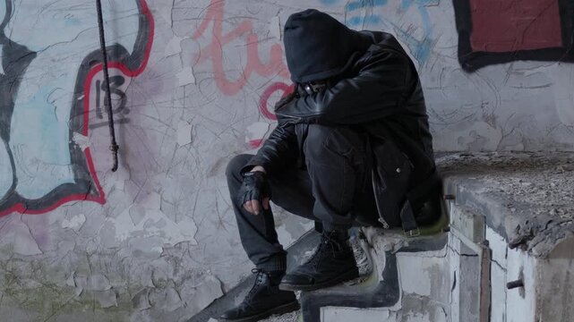 Hooded figure sits alone inside a graffiti-covered abandoned factory, creating a scene of mystery and urban decay, static tripod shot