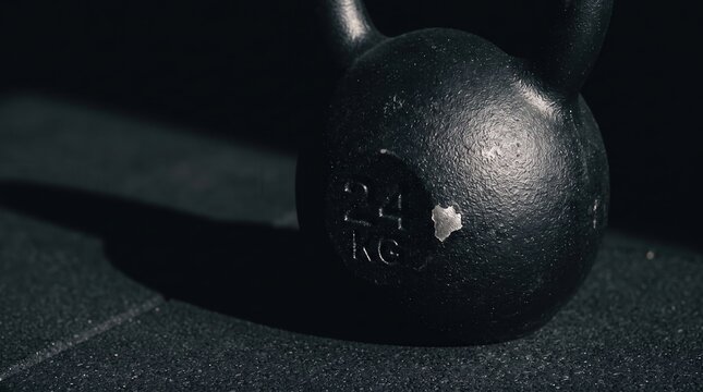 Black cast iron 24 kg kettlebell on a rubber gym floor with dramatic lighting, conveying strength training and powerlifting intensity for cross-training equipment marketing and fitness editorial.
