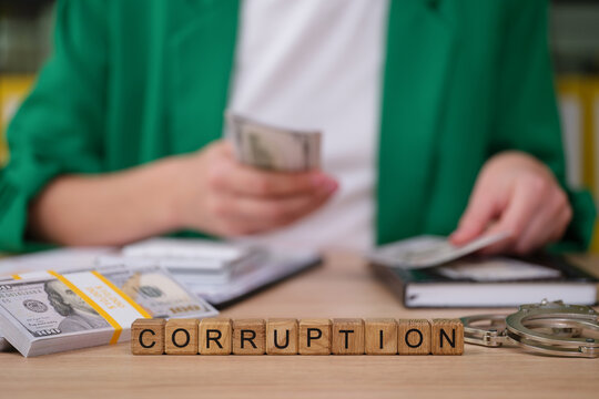 Stacks of cash sit on a desk alongside handcuffs and notebooks, as a person counts more money, illustrating the theme of corruption and financial misconduct in a corporate environment.